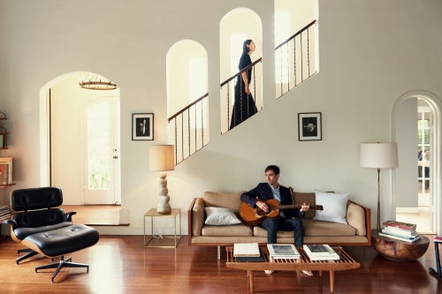Indoor shot of a man playing a guitar on a couch, with a woman on stairs in the background and modern decor in a brightly-lit living room.