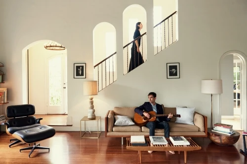 Indoor shot of a man playing a guitar on a couch, with a woman on stairs in the background and modern decor in a brightly-lit living room.