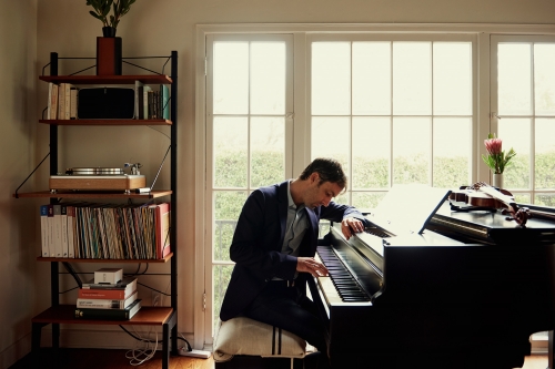 Man in a dark suit playing a grand piano indoors near a large multi-pane window with a shelving unit filled with records.
