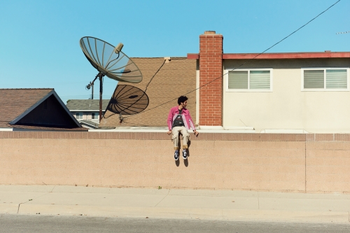 A person in a pink jacket sits on a wall in an urban setting, with a house, satellite dish, and blue sky in the background on a sunny day.