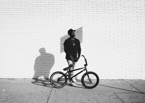 Monochrome shot of an African American man with a BMX bike against a white brick wall, showing urban style and athletic leisure.