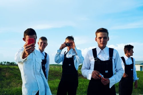 Group of five young men outdoors wearing traditional clothing, taking pictures with phones and camera under a blue sky.