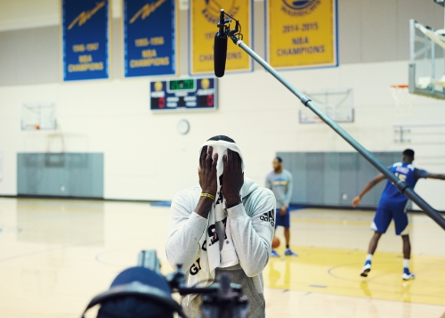 Basketball player with towel on head in gymnasium, standing in front of basketball court with teammates, recording equipment and banners.