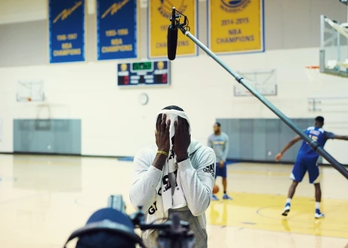 Basketball player with towel on head in gymnasium, standing in front of basketball court with teammates, recording equipment and banners.