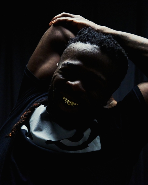Adult male smiles with gold teeth while posing in the dark with arms behind his head, showcasing a t-shirt with a white graphic design.
