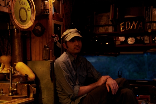 Man sitting in an indoor space with warm lighting, dressed casually with a cap, with rustic decorations and objects in the background.