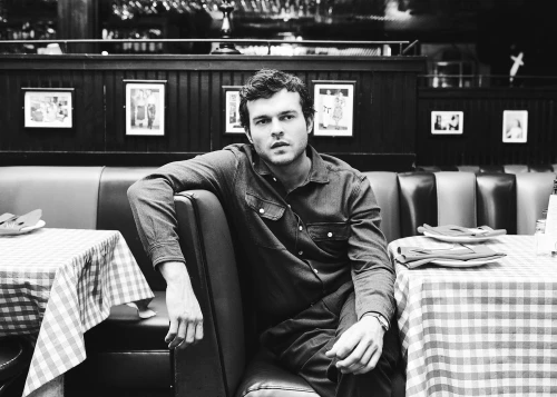 Monochrome portrait of a man sitting in a booth at an indoor restaurant, wearing a denim shirt with checkered tableclothes.