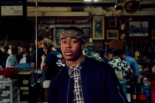 Portrait of a young adult man wearing a patterned shirt, dark jacket, and bandana in a barber shop with blurred patrons in background.