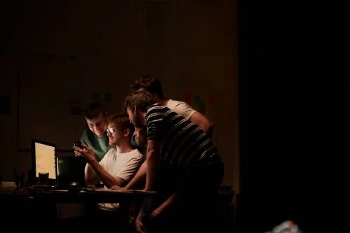 Five people gathered around a computer monitor in a dimly lit room, one person holds a cell phone, creating a collaborative atmosphere.