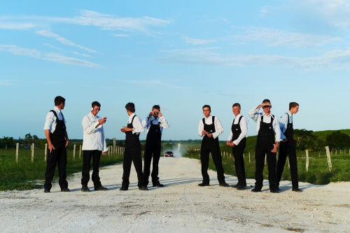 Seven men in rural setting wearing matching outfits with sky and a dirt road, including a car in the distance.