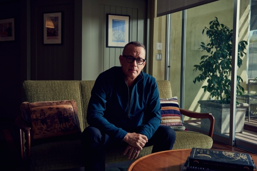 Mature man with glasses, in a blue top, sits on an olive couch indoors near a window, with a potted plant on the outdoor balcony.