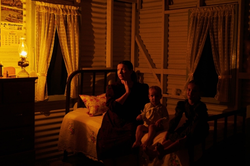 Woman and two children sitting on bed by a window in dimly lit room with yellow walls, bedside table and an oil lamp.