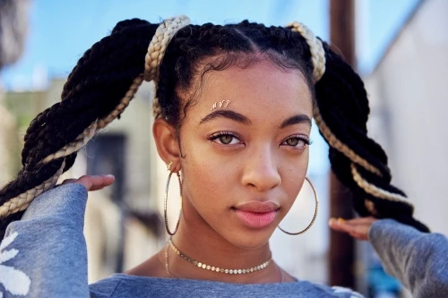 Close-up portrait of a young Black woman with braids, hoop earrings, and a choker necklace, set against a blurred outdoor background.
