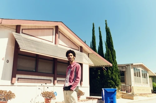 Man in patterned jacket stands outside a beige mobile home on a sunny day, posing casually with hand in pocket against a blue sky.