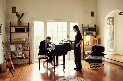 Indoor scene captures a man playing a piano while a woman stands nearby in a naturally lit, elegant home with hardwood floors.