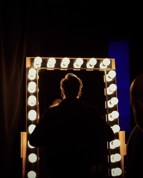 Back view of an adult looking in a lit vanity mirror in a dark room, possibly backstage, with a bright glow from the mirror.