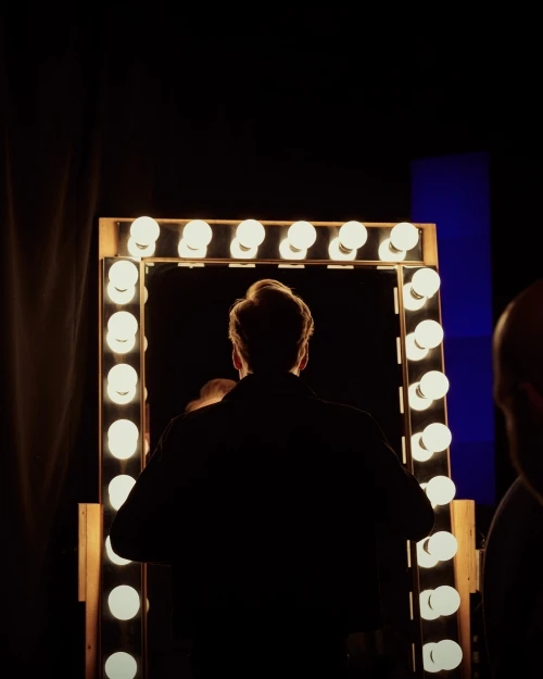 Back view of an adult looking in a lit vanity mirror in a dark room, possibly backstage, with a bright glow from the mirror.