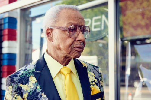 Elderly Black man in a floral jacket and yellow shirt stands outside a shop with an OPEN sign in the window, looking off to the side.