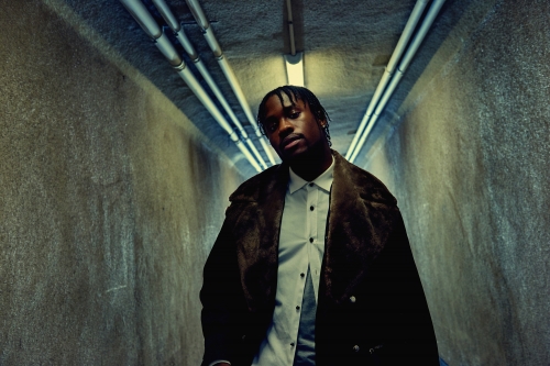 Portrait of an adult male wearing a fur coat and white shirt in an indoor tunnel environment with overhead lighting.