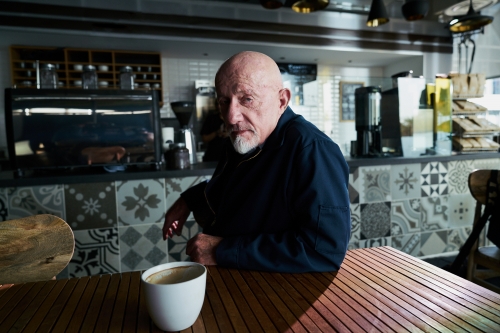 An older man with a serious expression sits at a wooden table in a cafe, with a coffee cup in front of him and an espresso machine in the background.