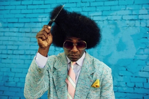 Adult male with afro hairstyle, wearing a blazer and sunglasses, is holding a comb near his head in front of a brick wall.