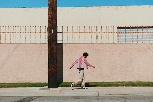 A person walks beside a wall with a rusted fence, set against a blue sky, with a wooden utility pole in the foreground.