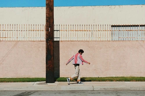 A person walks beside a wall with a rusted fence, set against a blue sky, with a wooden utility pole in the foreground.