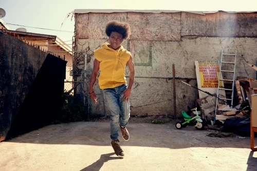 Young adult in casual wear walks toward the camera in an outdoor urban setting with a weathered wall backdrop, capturing a candid moment.