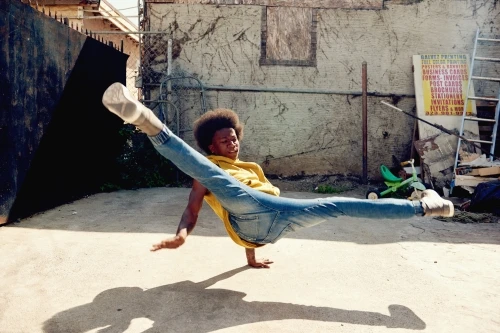Young adult male breakdancer performing a split in an outdoor urban setting, wearing jeans and a yellow shirt, with a concrete ground.
