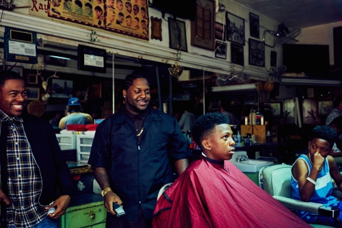 Barbershop scene shows three men and a young customer with a red cape getting a haircut. Walls are decorated with various photos.