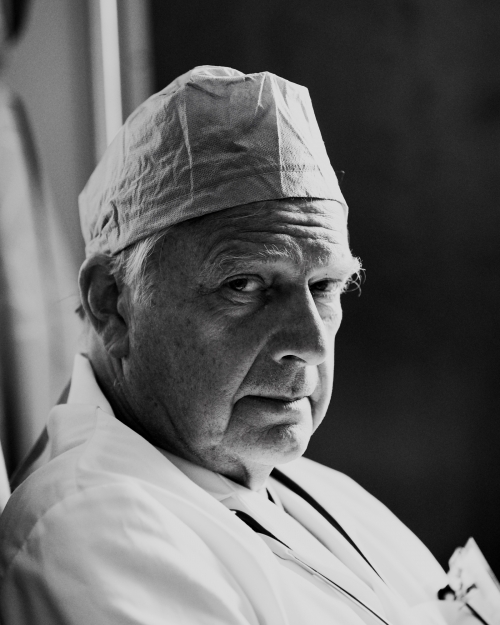 Black and white portrait of a mature adult male wearing a surgical cap and white coat, captured in an indoor medical setting.