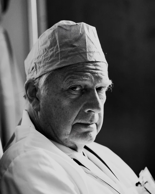 Black and white portrait of a mature adult male wearing a surgical cap and white coat, captured in an indoor medical setting.