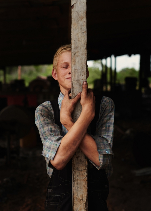 Young man in overalls embraces weathered wooden post indoors, bathed in soft light, with muted colors creating a rural atmosphere.