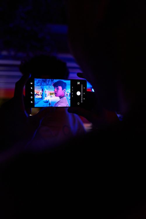 A man holds a smartphone displaying an image of a young man in a brightly lit setting, all against a dark, shadowed background.