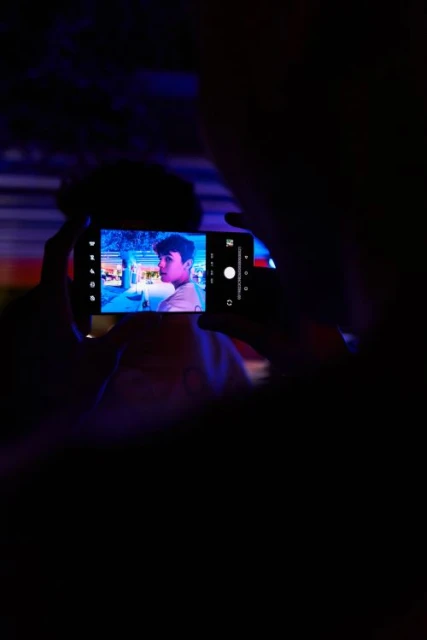 A man holds a smartphone displaying an image of a young man in a brightly lit setting, all against a dark, shadowed background.