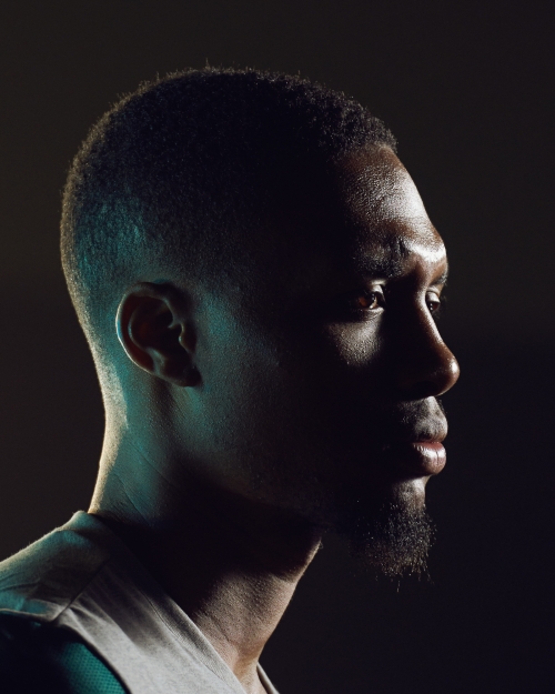 Profile of an African American man with short hair and a goatee, gazing off into the distance, illuminated by blue and yellow lighting.