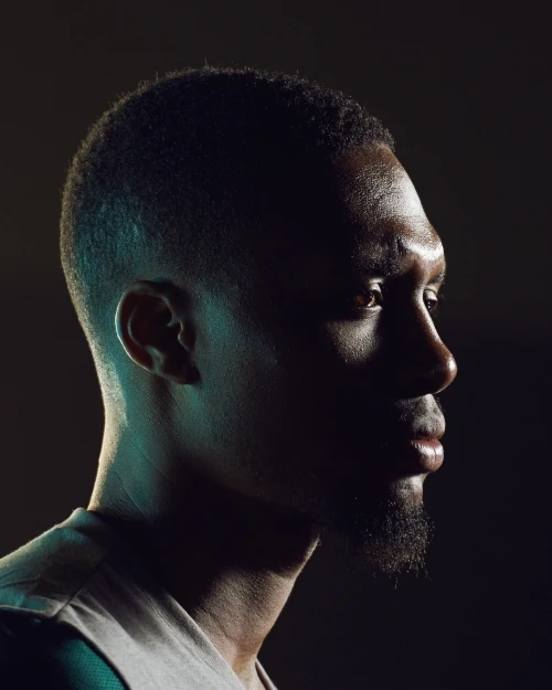 Profile of an African American man with short hair and a goatee, gazing off into the distance, illuminated by blue and yellow lighting.