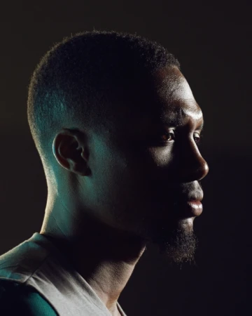Profile of an African American man with short hair and a goatee, gazing off into the distance, illuminated by blue and yellow lighting.