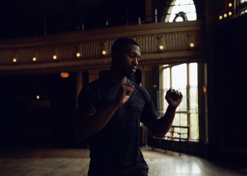 Athletic African-American man in activewear stands poised in a dark gymnasium with vintage architecture and arched windows, looking down.