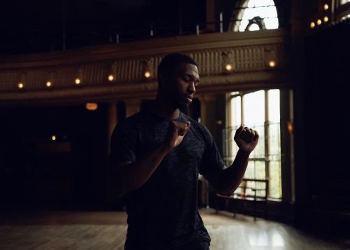 Athletic African-American man in activewear stands poised in a dark gymnasium with vintage architecture and arched windows, looking down.