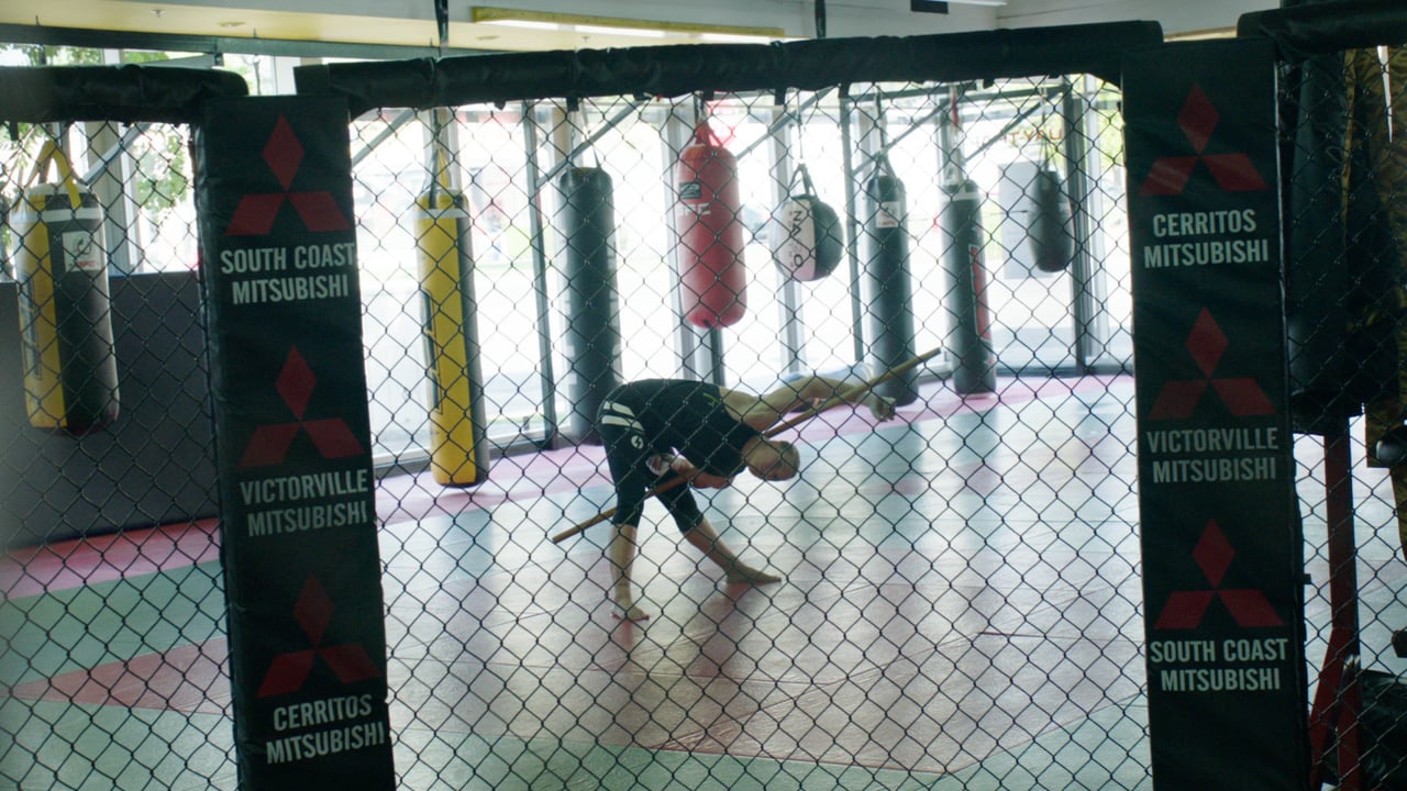 Man stretches with a wooden stick inside a caged mma fighting area with punching bags hanging in background and advertisements on corner posts.