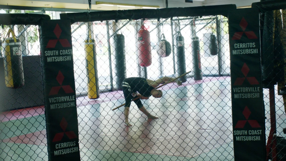Man stretches with a wooden stick inside a caged mma fighting area with punching bags hanging in background and advertisements on corner posts.