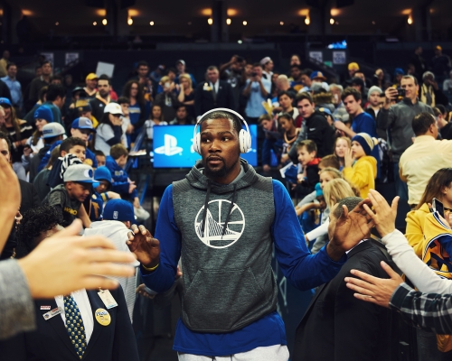 Basketball player in a grey hoodie and headphones walking through a crowd, indoor arena with bright overhead lighting.