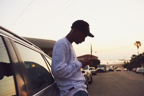 Young man in a baseball cap leans against a car, using a smartphone on a street with other parked cars, silhouetted against an evening sky.