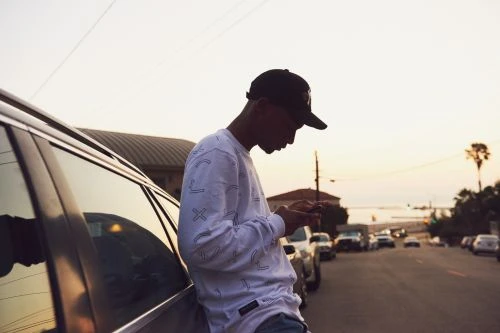Young man in a baseball cap leans against a car, using a smartphone on a street with other parked cars, silhouetted against an evening sky.