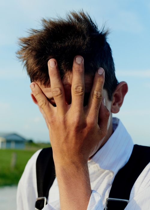 Close-up of a fair-skinned young adult with dark hair, covering their face with their hand against a rural landscape and blue sky.