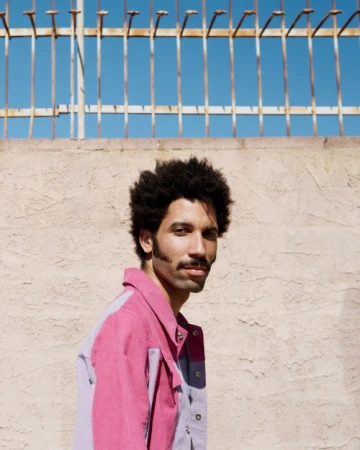Man with afro and mustache wearing a pink corduroy jacket against a stucco wall and blue sky