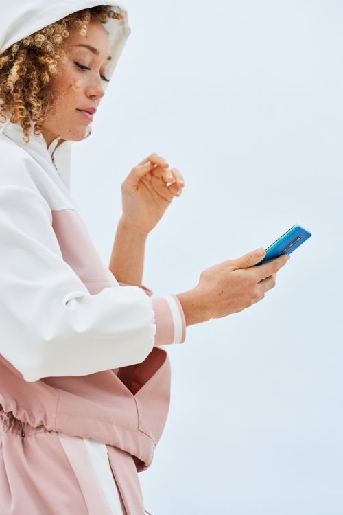 Woman in a pink and white hooded jacket holding a blue smartphone against a seamless white background.