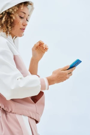 Woman in a pink and white hooded jacket holding a blue smartphone against a seamless white background.