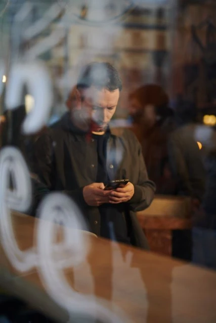 Man checks his phone in front of a blurred background, with reflective writing on glass, creating a hazy, urban, night scene.